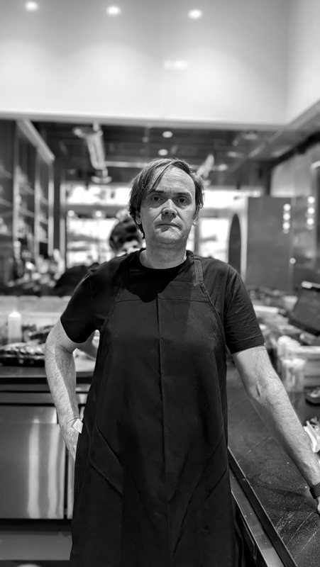 Torsten Schulz in the kitchen at L Campo, black apron, the dining room behind him.