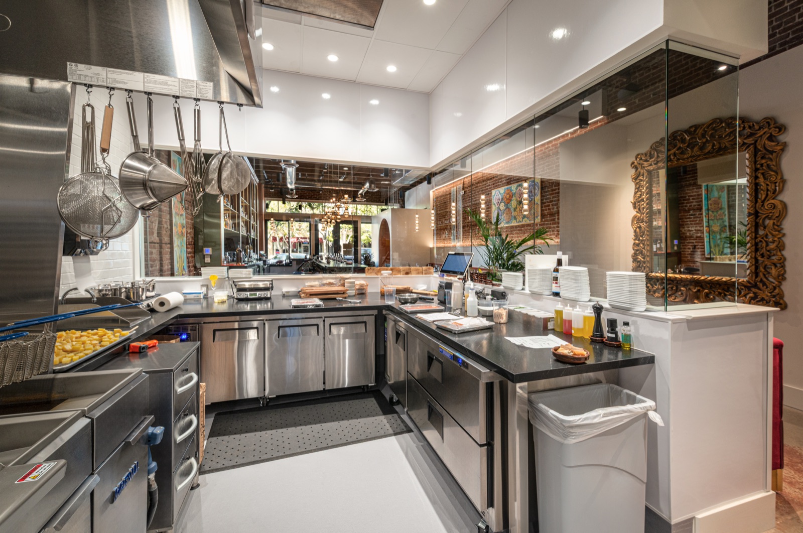 The open kitchen at L Campo: a black stone counter in a U, steel drawers and low refrigerators beneath, whisks and strainers under the hood, and through the glass the dining room, the brick and the street.