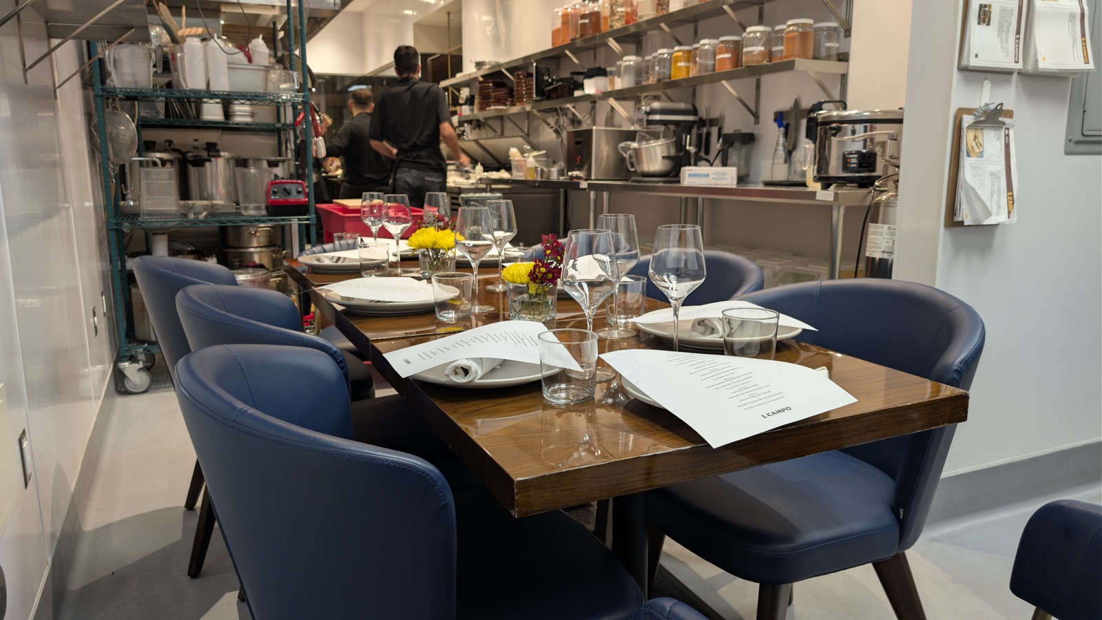 Six blue chairs at a wooden table set with glasses and printed menus, in the working kitchen; two cooks at the range behind, spice jars on the shelves above.