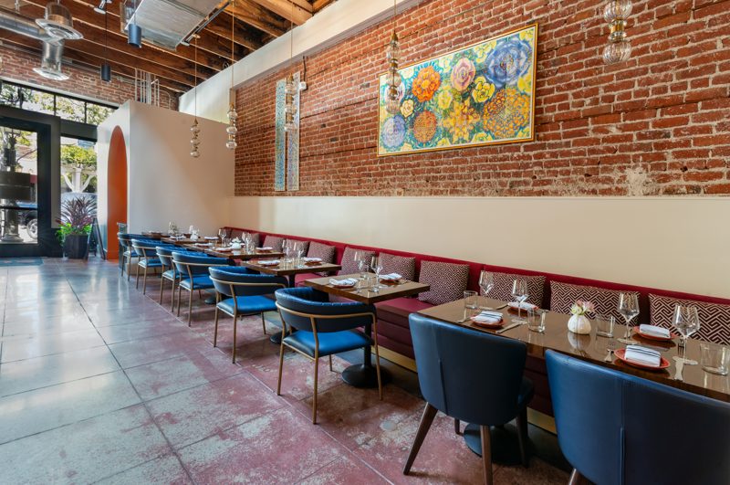 The dining room at L Campo: blue chairs and a long red banquette along the brick wall, tables set, a painting of flowers above.