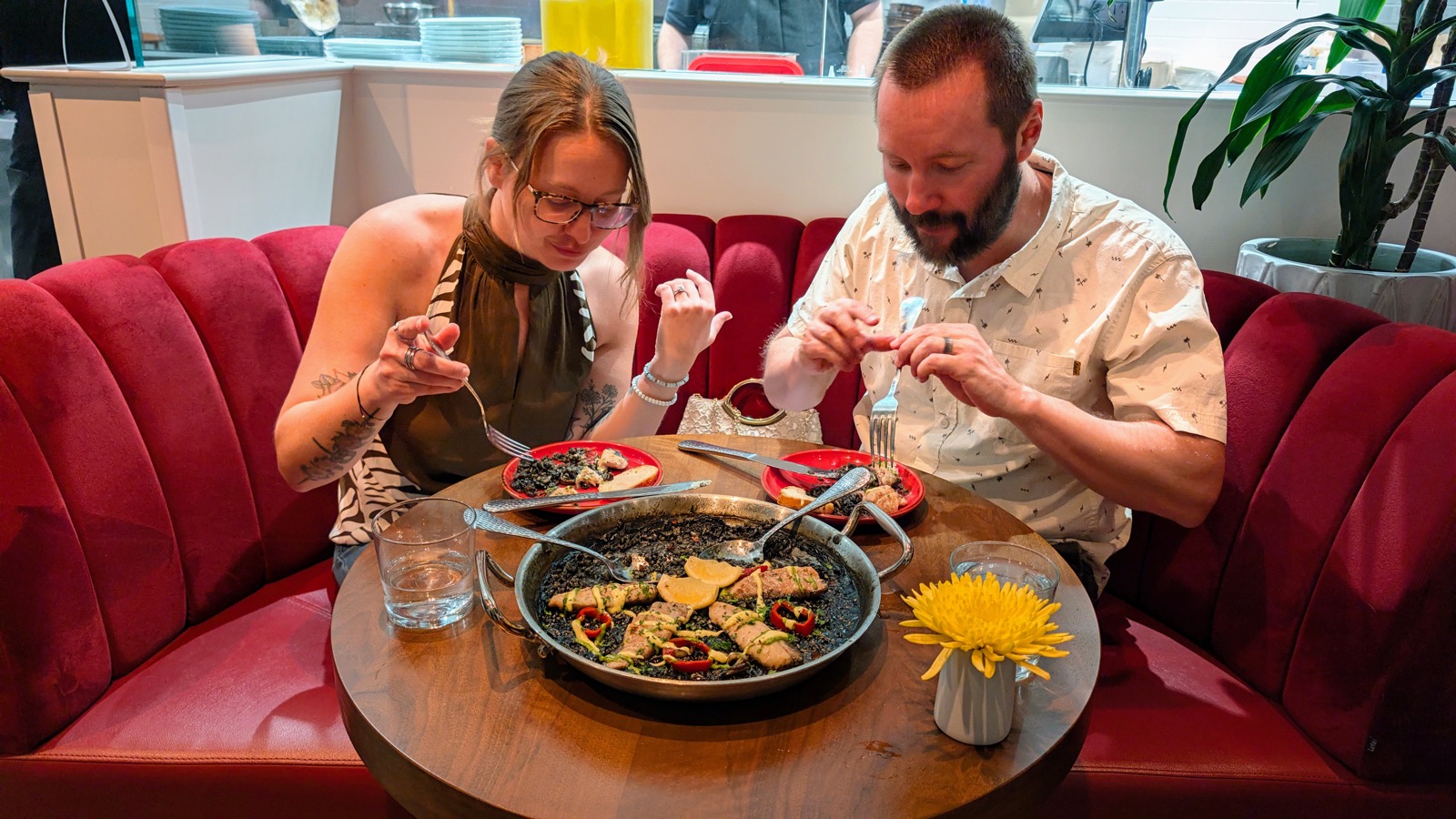 Rose and Casey at a round wooden table in a red velvet booth, heads bent over a black paella in its pan; the open kitchen and stacks of plates behind them, a yellow chrysanthemum in a small vase.