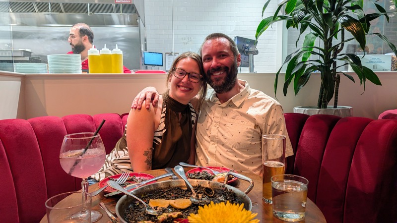 Rose and Casey, smiling at the camera with their arms around each other, the emptied paella pan and a pink gin tonic in front of them, a cook at the window behind.