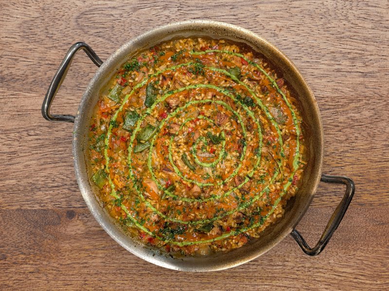 The Valenciana from above on a wooden table: rice, rabbit and green beans under spirals of mojo verde and mojo picón, parsley scattered.